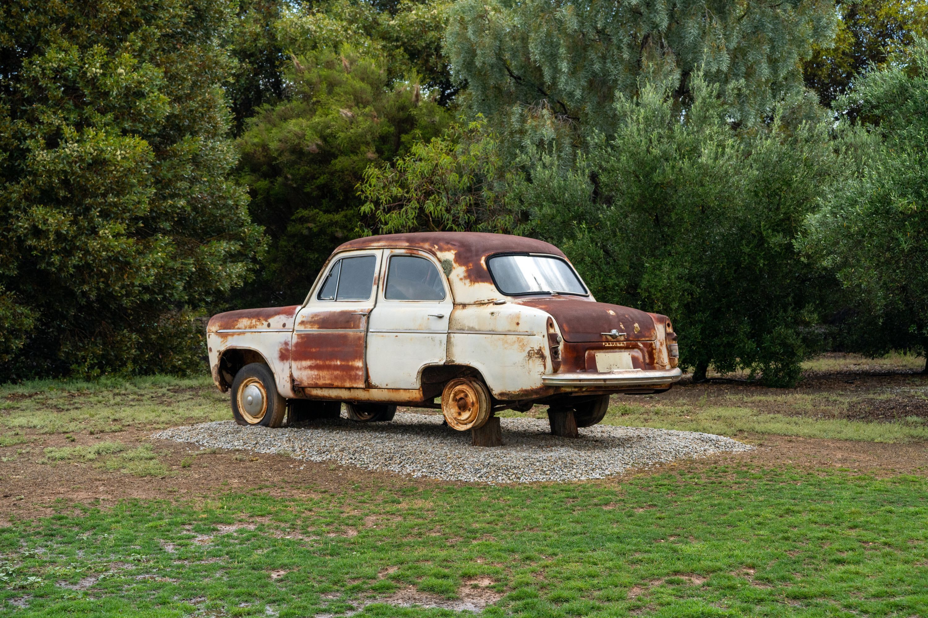 An old, rusted car parked on a gravel area surrounded by grass and trees.