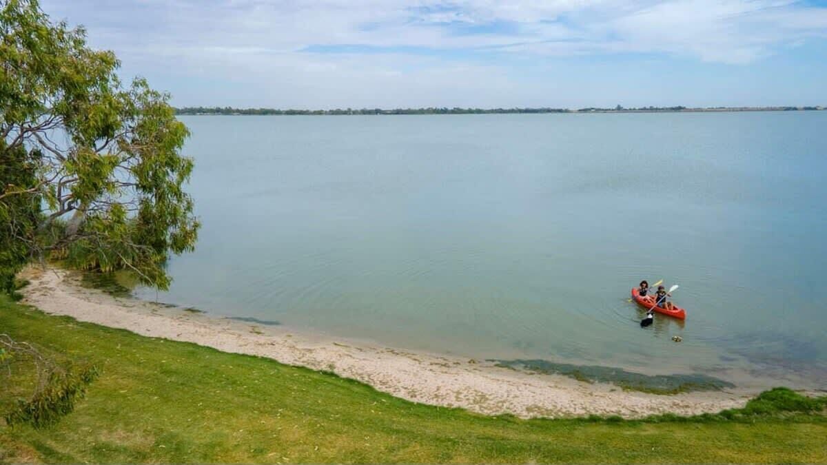 Un lac paisible, avec une plage de sable et un arbre sur la gauche. Un kayak rouge, avec deux personnes à bord, est sur l’eau.