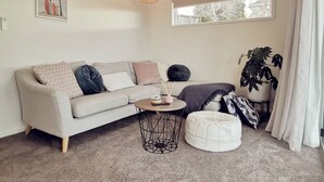 A modern living room with a sofa, round coffee table, and a pouf. There is a window with a view, pendant lights, and an air conditioning unit on the wall. - Huntsbury Hideaway, Cottage with amazing views (Christchurch)