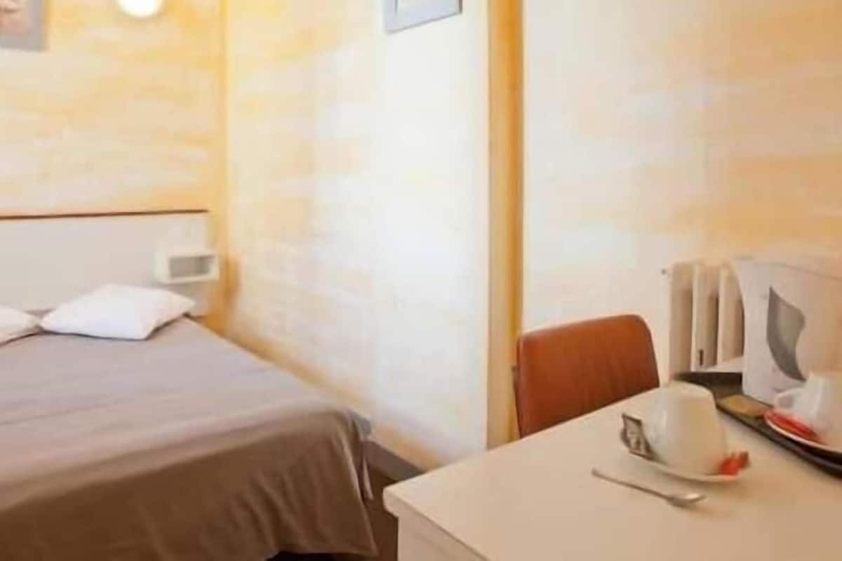 A hotel room with a bed, a table with a cup and saucer, and a chair.