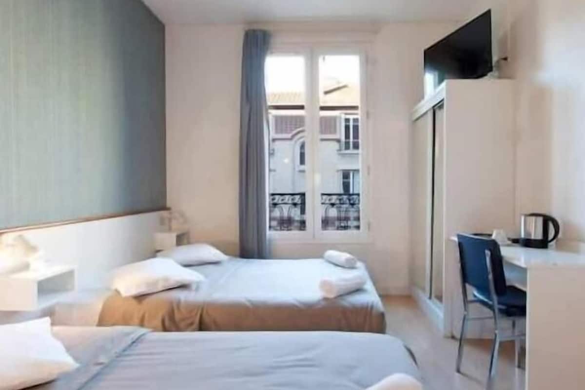 A hotel room with two beds, a desk, a chair, and a window with a view of buildings.