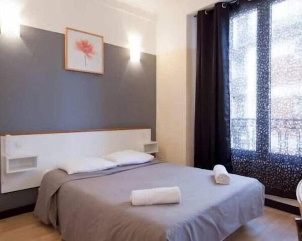 Habitación de hotel con cama, cabecera, un cuadro enmarco, ventana con cortinas y una toalla sobre la cama.