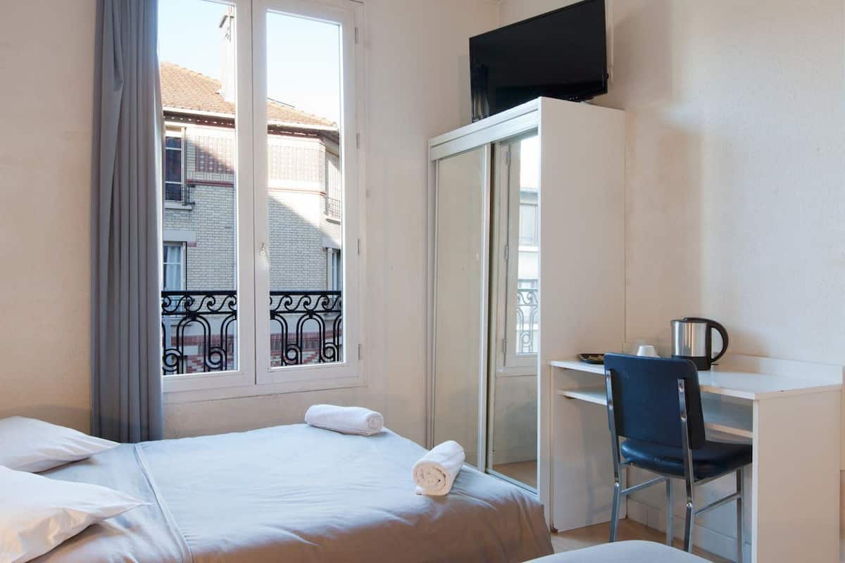 A hotel room with a bed, a desk with a chair, a television, and a balcony with a view of buildings.