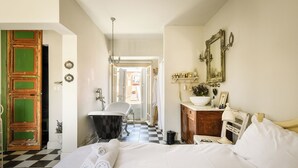 Une chambre d’hôtel avec un sol à carreaux, un lit recouvert de draps blancs, une baignoire ancienne, une commode en bois et un miroir au mur.