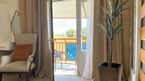 Una habitación con una silla, una planta en maceta y vistas a un balcón con una valla azul y un coche rojo.