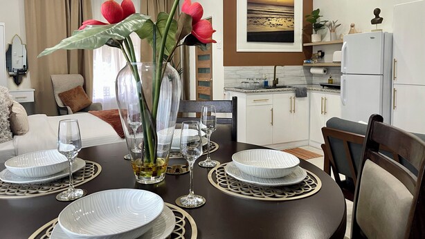 Un comedor con una mesa redonda dispuesta para cuatro personas, rodeada de sillas. Hay un florero con flores sobre la mesa, y un cuadro de un atardecer en la pared detrás de la mesa.