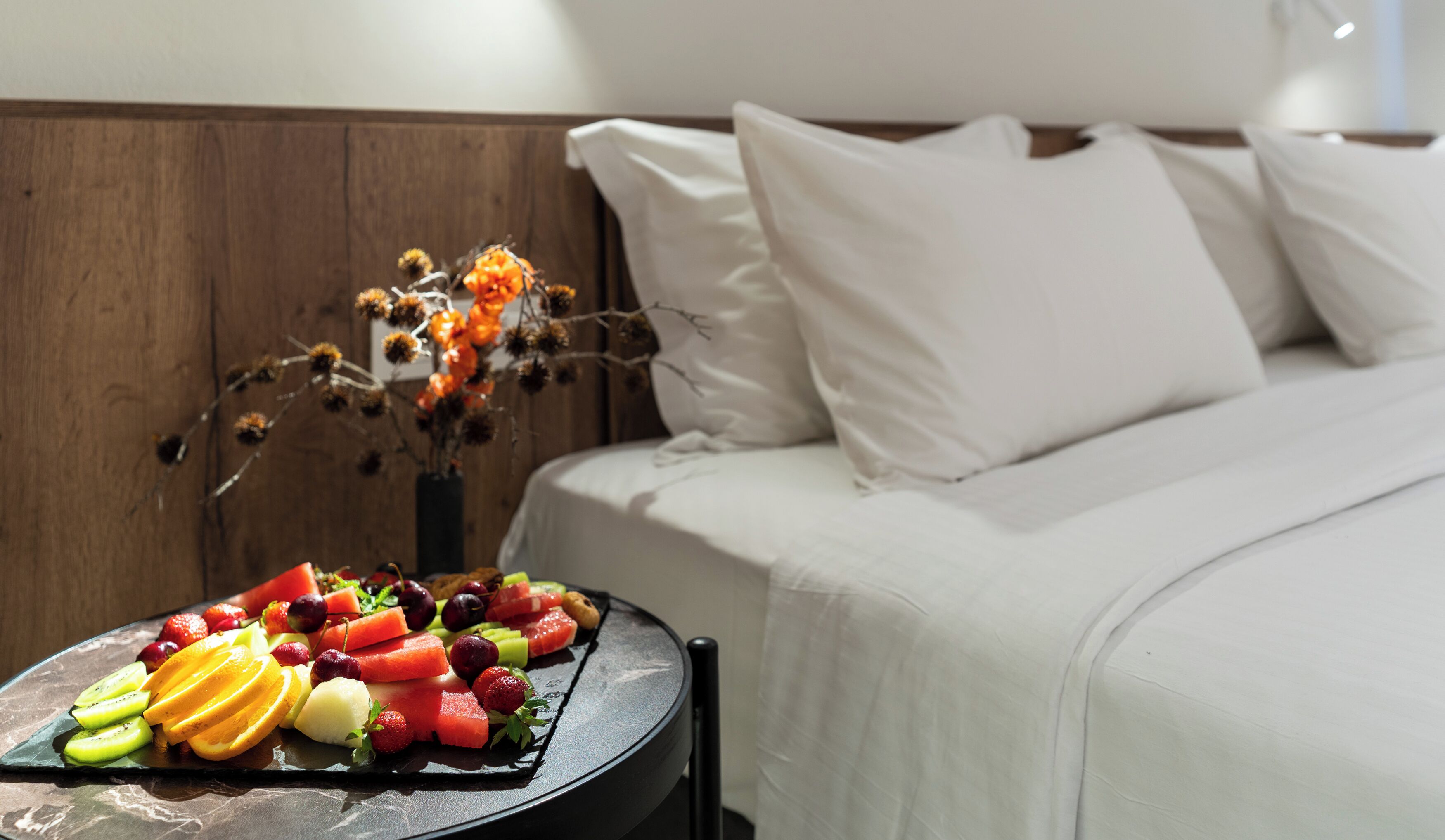 Una cama bien hecha con sábanas blancas y una bandeja de frutas sobre una mesita auxiliar.