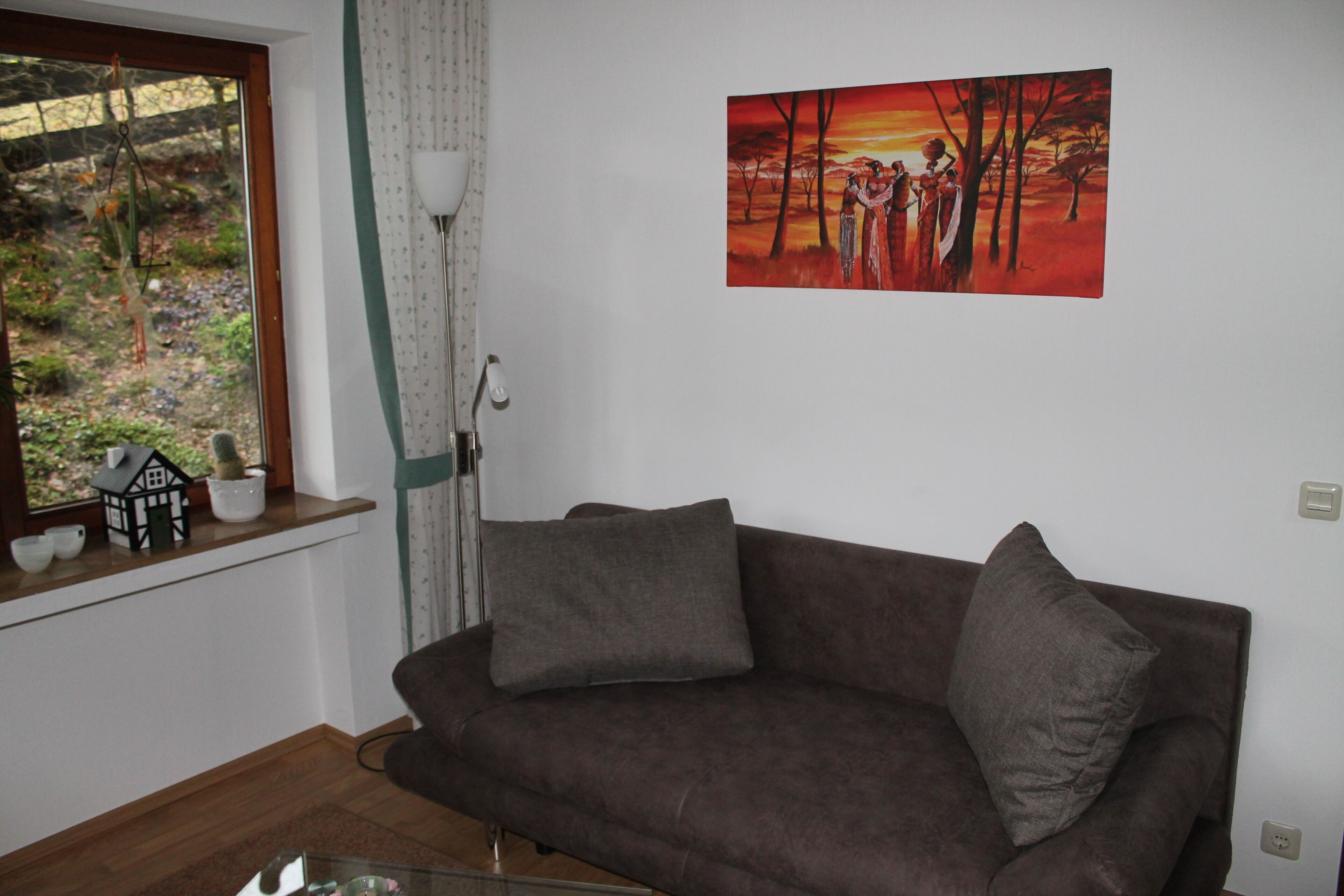 Un salon avec un canapé marron, une fenêtre donnant sur un paysage verdoyant et un tableau accroché au mur.