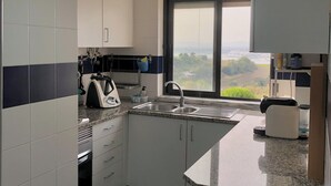 A modern kitchen with white cabinets, a granite countertop, a built-in oven, a kettle, and a view through a large window. - Duplex in Caparica (Caparica)