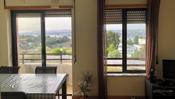A room with a balcony, a table with a checkered tablecloth, a potted plant, and a TV stand with electronic devices. - Duplex in Caparica (Caparica)