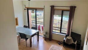 A room with a dining table, chairs, a sofa, a small table, a TV, and a window with curtains. - Duplex in Caparica (Caparica)