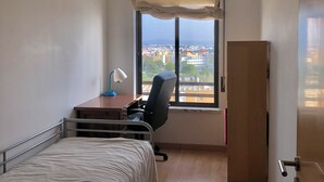A bedroom with a bed, desk, chair, and a window with a view of a cityscape. - Duplex in Caparica (Caparica)