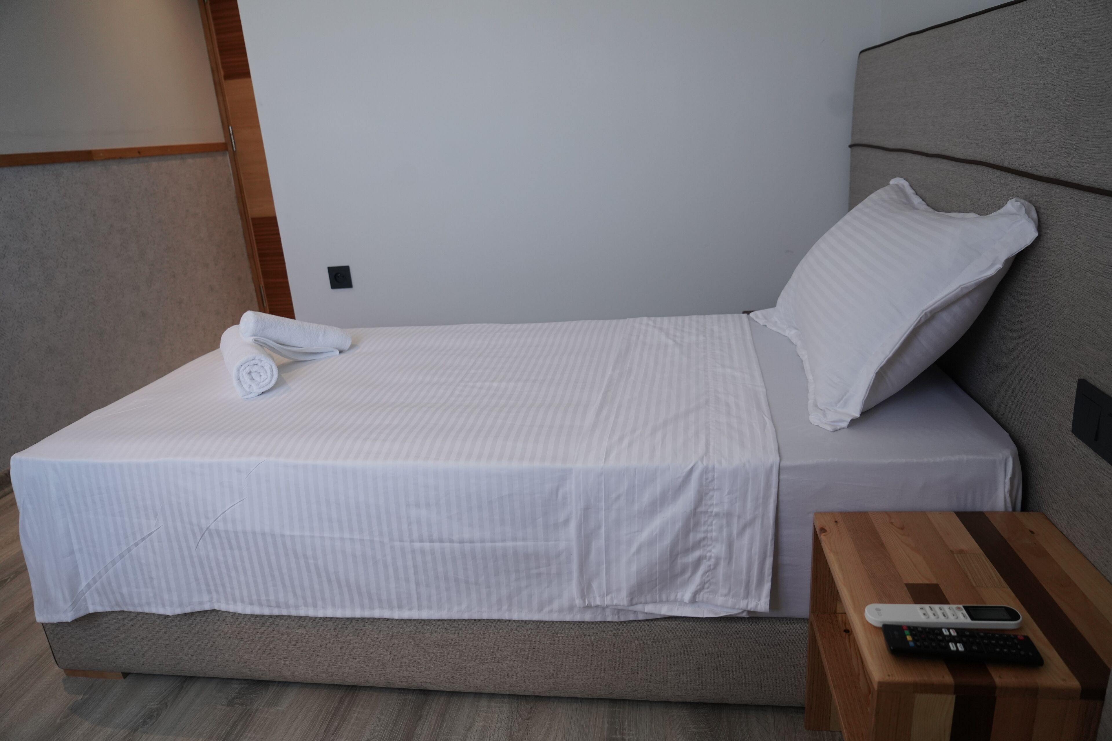 Una cama bien hecha con cubrecama y almohadas blancas, una mesita de noche de madera con un control remoto y una lámpara de pared.