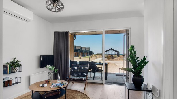 Ein modernes Wohnzimmer mit Fernseher, Esstisch und Sofa. Ein großes Fenster bietet Blick auf ein Gebäude und einen Strand.
