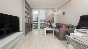 A modern living room with a flat-screen TV, grey sofa, white coffee table, and a balcony with a view.