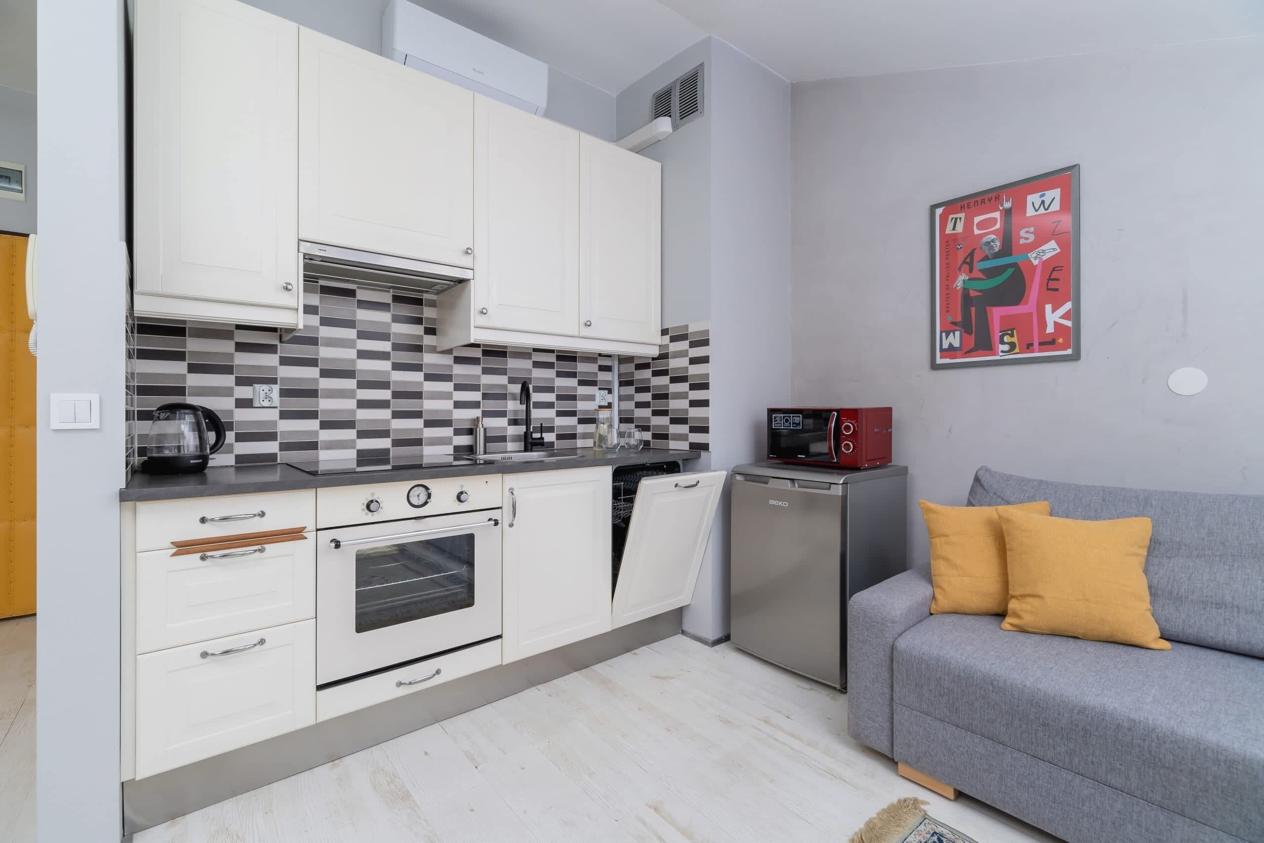 A compact kitchen with white cabinets, a dishwasher, a microwave, and a small oven. There is a grey sofa with yellow cushions in the adjacent living area.