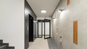 Un pasillo moderno con una escalera a la izquierda, una puerta de ascensor de vidrio en el centro y un panel de madera decorativo en la pared.