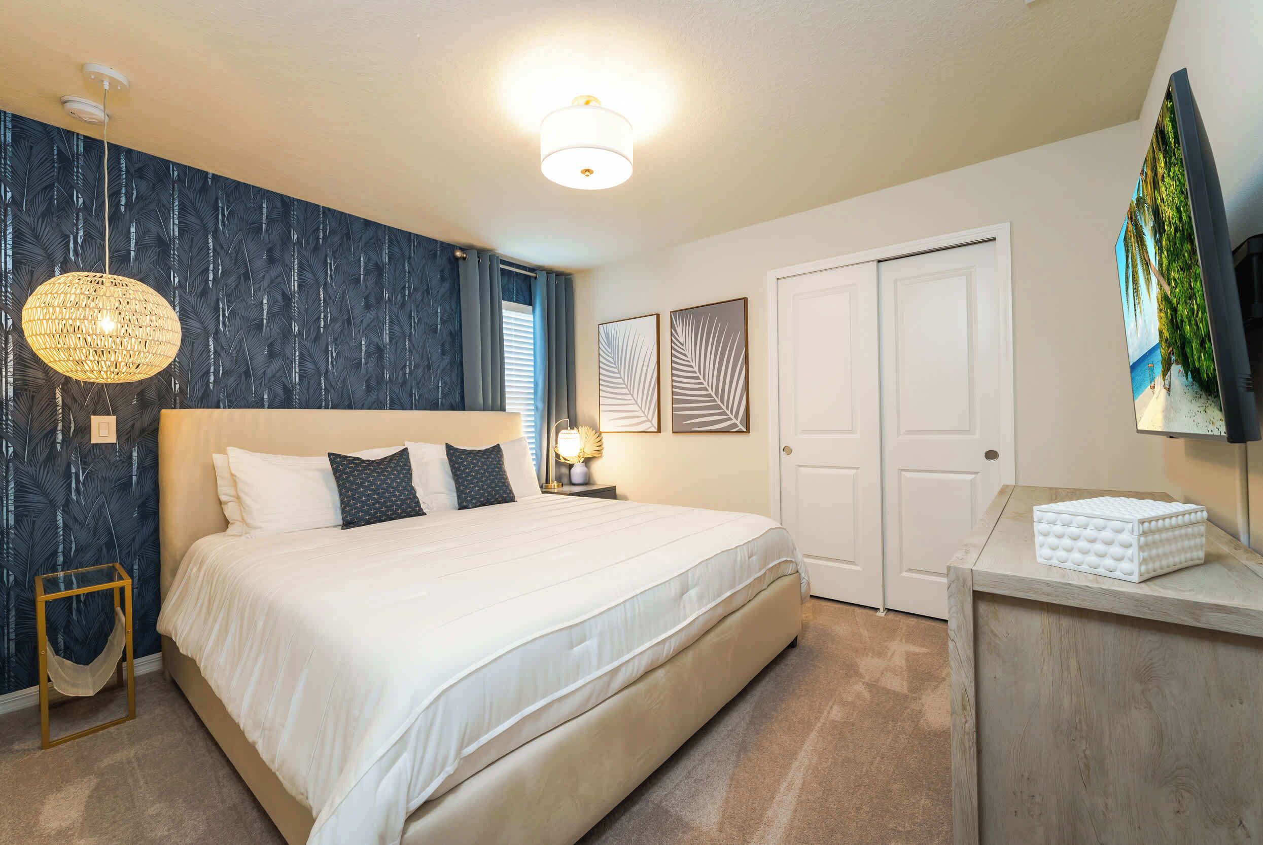 A bedroom with a large bed, a bedside table with a lamp, a wall-mounted TV, and a door.