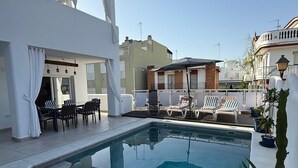 En pool på taket med solstolar och ett bord, omgiven av vita väggar och en klar himmel.