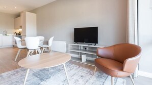 Un salon moderne avec un téléviseur à écran plat, un canapé en cuir marron, des chaises blanches et une table basse en bois.