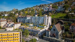 Un hotel de varios pisos con diseño moderno, situado en una zona montañosa con edificios residenciales al fondo.
