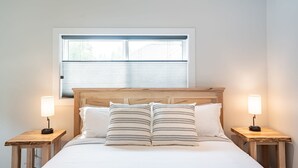 A bedroom with a wooden headboard, a window with blinds, two bedside tables with lamps, and a bed with white bedding and striped pillows.