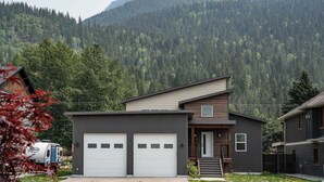 A modern two-story building with a garage, a small porch, and a view of a forested mountain.