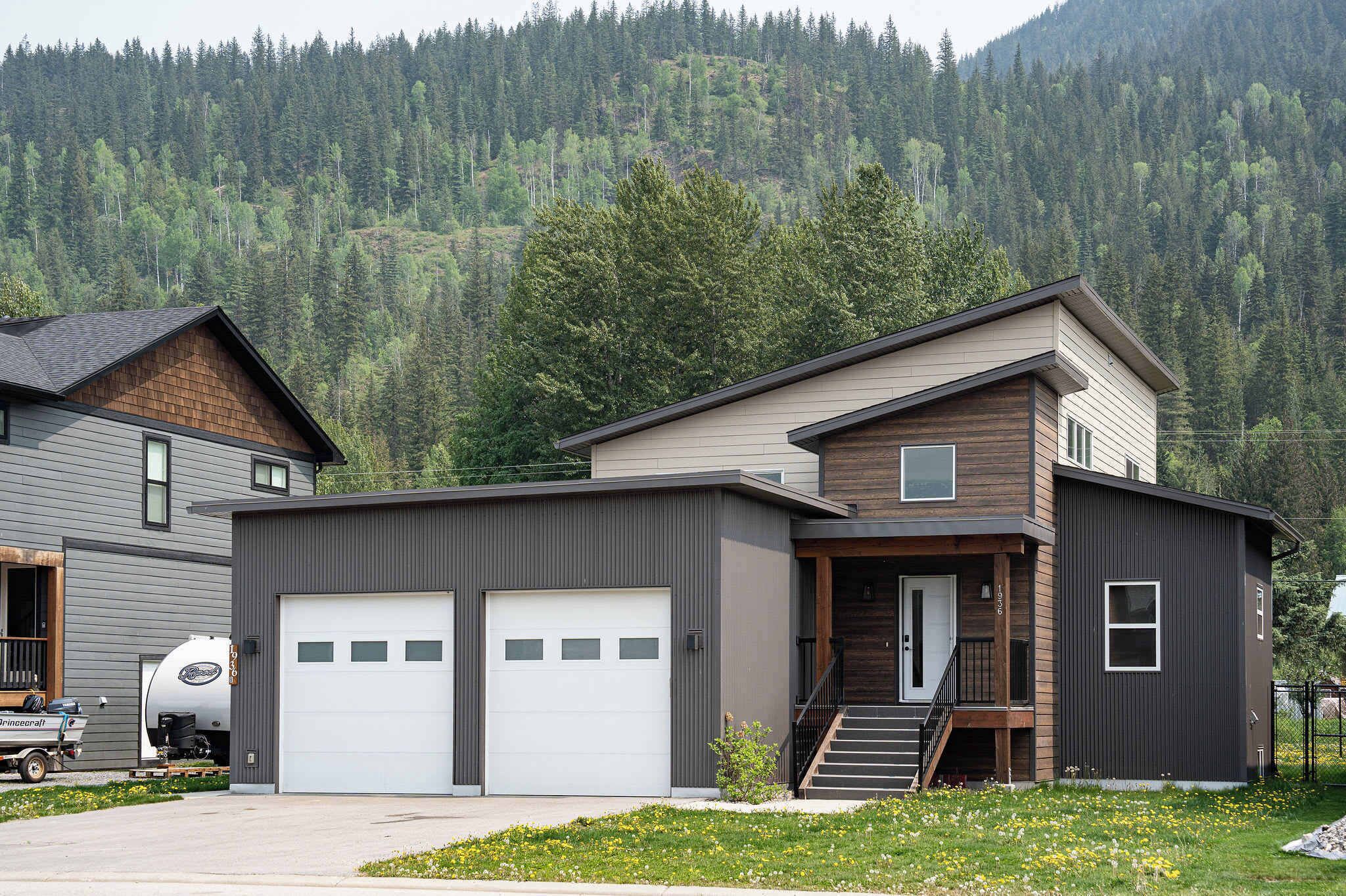 A modern two-story house with a garage, a staircase leading to the second floor, and a forested mountain backdrop.