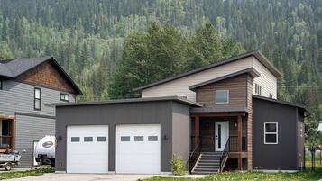 A modern two-story house with a garage, a staircase leading to the second floor, and a forested mountain backdrop.