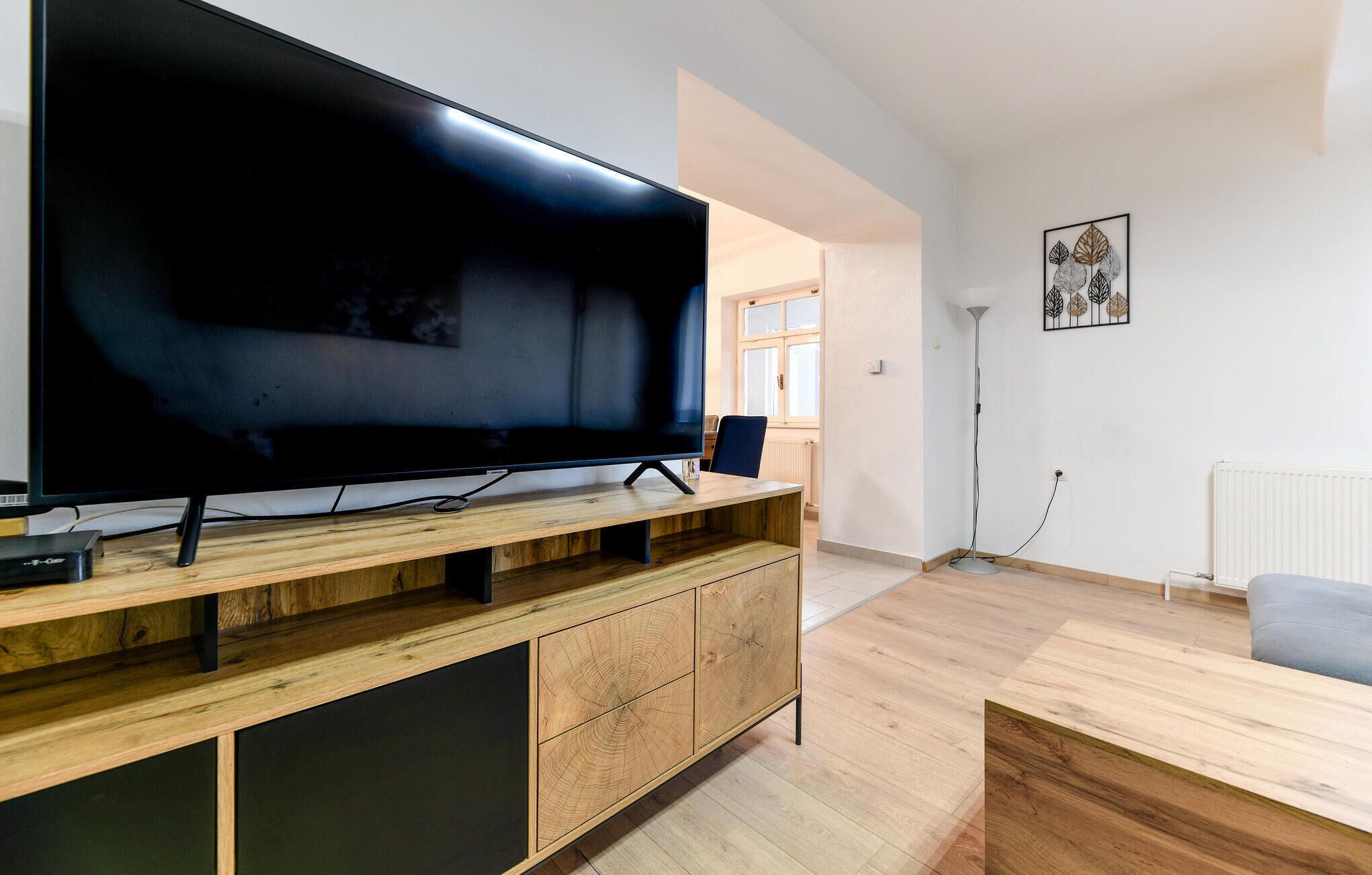 Una sala de estar moderna con un televisor de pantalla plana grande sobre un mueble de madera, una lámpara de pie y un sofá.