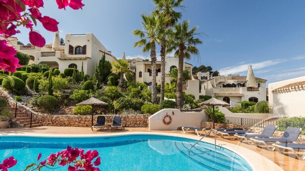 A swimming pool with lounge chairs, a stone wall, and a building with multiple balconies and palm trees. - La Manga Club Los Altos Townhouse 489 (Atamaría)