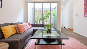 A modern living room with a glass coffee table, a gray sofa with colorful cushions, and a large window offering a view of greenery outside.