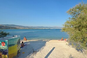 A beach with people sunbathing and swimming, a Coca-Cola cooler, and a clear view of the sea and distant hills.