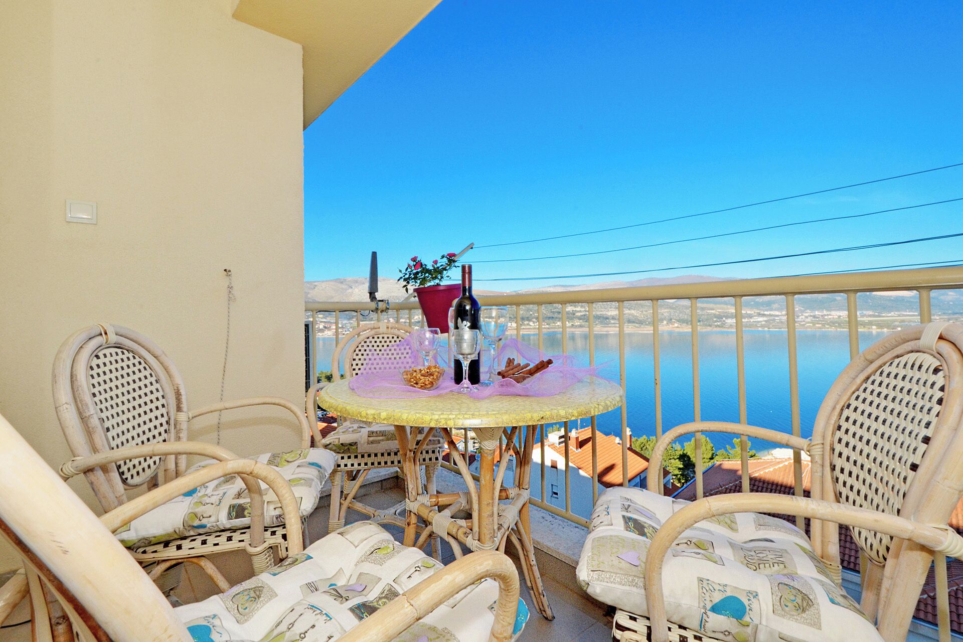 A balcony with wicker chairs and a round table, overlooking a body of water and a distant cityscape.