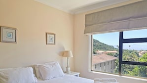 A hotel room with a bed, a nightstand with a lamp, and a window with a view of buildings and greenery. - Seahorse (Plettenberg Bay)