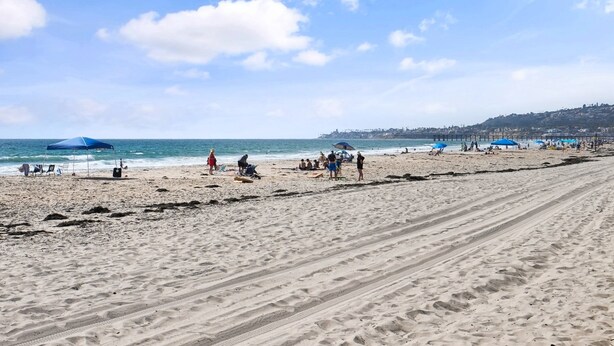 A beach with people, umbrellas, and a pier in the background. - Mission Bay Condo - Steps to Sand, Dining, AC, Parking, Location & Shops! (San Diego)