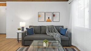 A modern living room with a grey sofa, a glass coffee table, and a small side table with a lamp. There are three framed artworks on the wall above the sofa. - Mission Bay Condo - Steps to Sand, Dining, AC, Parking, Location & Shops! (San Diego)