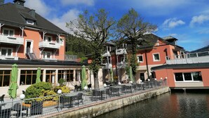 A riverside hotel with balconies, outdoor seating, and a canal. - Strandhotel Burgstaller (Feld am See)