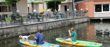 Two people on paddleboards in a canal with a restaurant and outdoor seating in the background.