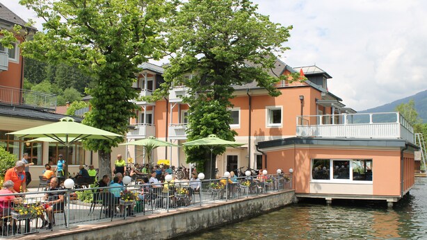 A riverside dining area with outdoor seating, a boat house, and a view of the water. - Strandhotel Burgstaller (Feld am See)