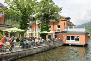 A riverside dining area with outdoor seating, a boat house, and a view of the water. - Strandhotel Burgstaller (Feld am See)