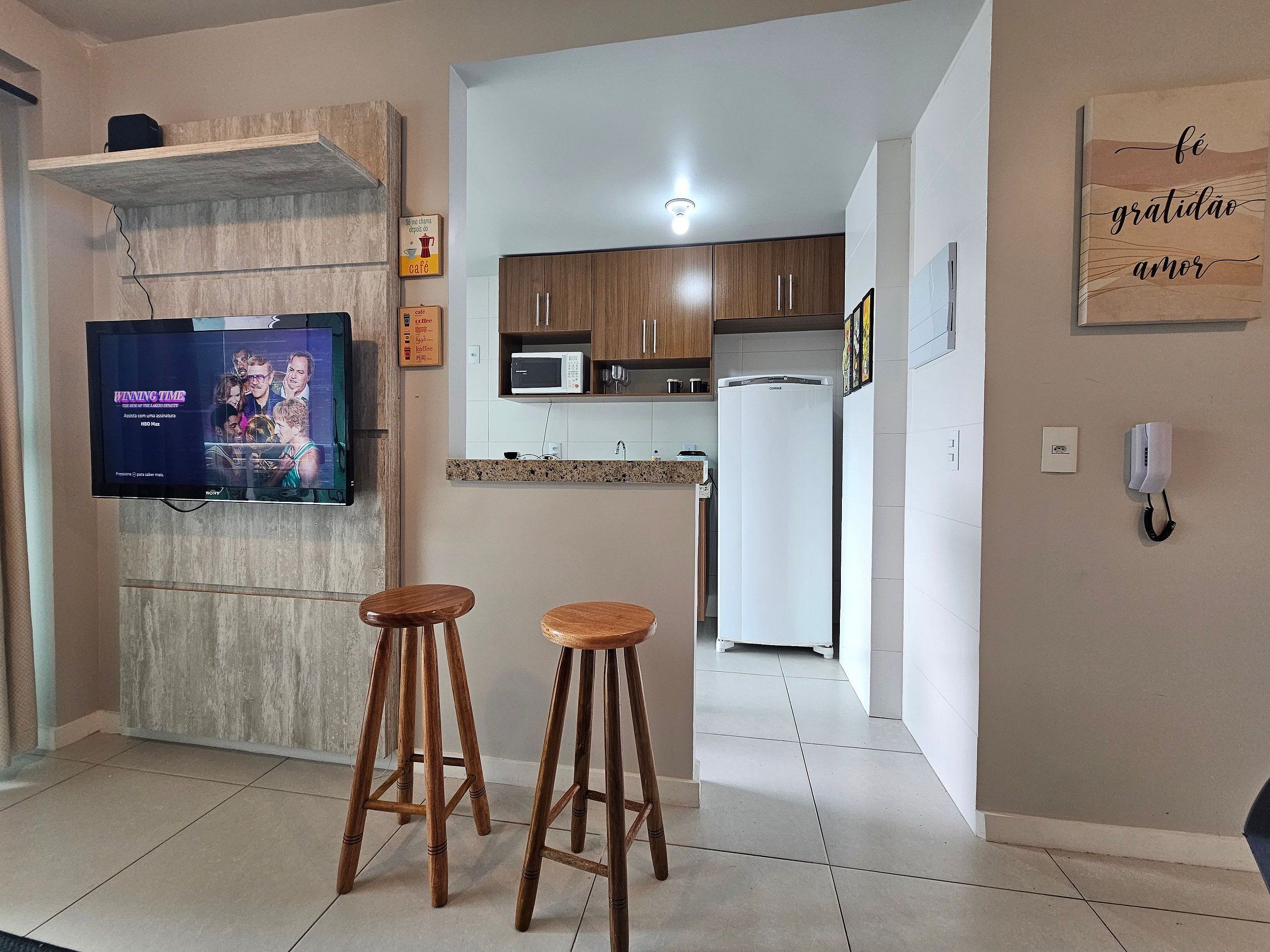 A modern kitchen with a flat-screen TV, wooden stools, and a refrigerator.