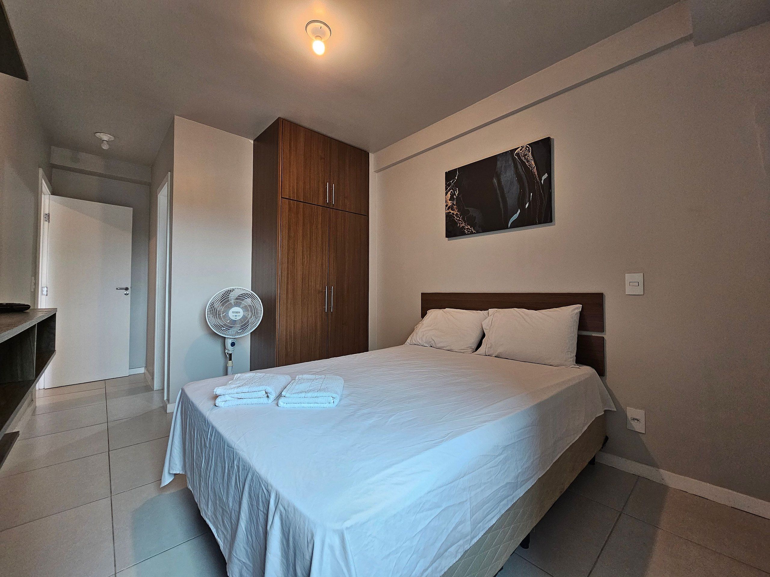 A bedroom with a bed, a wardrobe, a fan, and a painting on the wall.