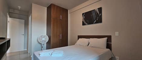 A bedroom with a bed, a wardrobe, a fan, and a painting on the wall.