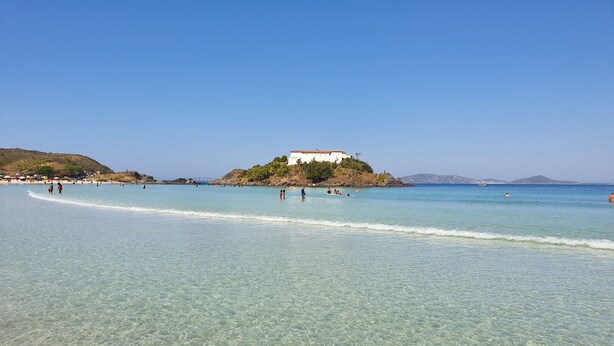 A beach with clear shallow waters, a white building on a hill, and a few people in the water. - Lindo AP Praia do Forte - Porto Azul 106 (Cabo Frio)
