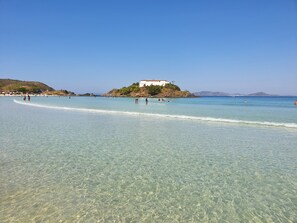 A beach with clear shallow waters, a white building on a hill, and a few people in the water. - Lindo AP Praia do Forte - Porto Azul 106 (Cabo Frio)