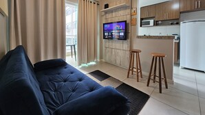 A living room with a blue sofa, wooden stools, a kitchen with a microwave and refrigerator, and a TV mounted on the wall. - Lindo AP Praia do Forte - Porto Azul 106 (Cabo Frio)