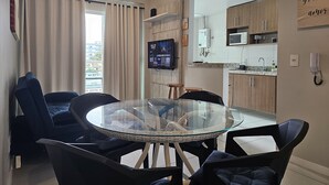 A modern kitchen with a microwave, sink, and wooden cabinets. There's a glass table with chairs and a TV mounted on the wall. - Lindo AP Praia do Forte - Porto Azul 106 (Cabo Frio)