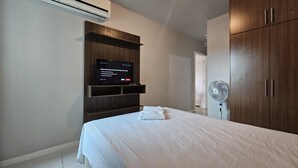 A bedroom with a bed, a TV mounted on a wooden shelf, an air conditioner, and a fan. - Lindo AP Praia do Forte - Porto Azul 106 (Cabo Frio)