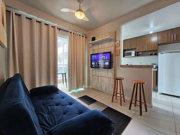 Una sala de estar con un sofá azul, un ventilador de techo, un televisor de pantalla plana montado en la pared, muebles de cocina de madera y un refrigerador.